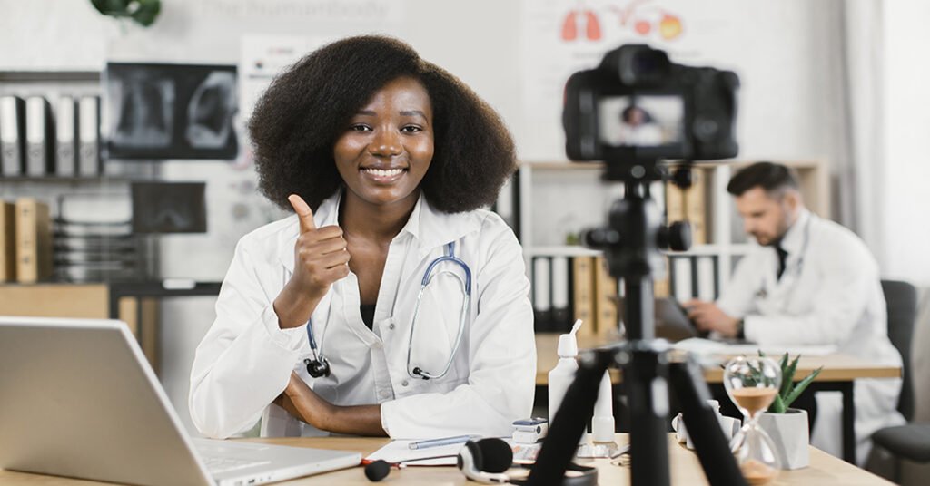 Smiling medical staff wellness staff woman in lab coat sitting at desk with modern laptop and using camera for recording video blog and showing thumb up. Qualified female doctor creating content for her medical channel.
